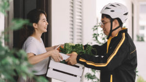 A young man with a bicycle helmet delivers a crate full of fresh vegetables to a woman standing at her front door