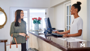 A traveler with her luggage is greeted by an enthusiastic front desk worker