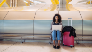 A woman sits next to her luggage at an airport, typing on her laptop