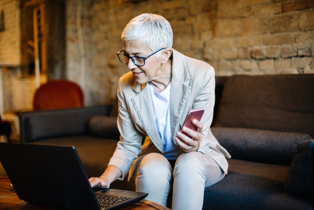 An lady with grey hair, glasses, and a pantsuit sits on a couch while working on her phone and laptop.
