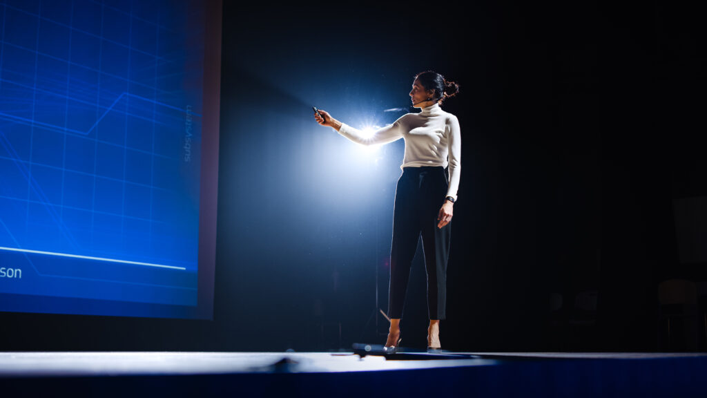 A woman in business attire stands in the spotlight on a stage while giving a presentation