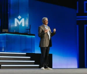 A man in a suit speaks on stage beside steps and a desk with a computer. The background is blue with a large M logo displayed on a screen.