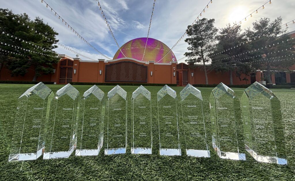 Nine glass awards are lined up on grass in front of an outdoor venue with a colorful, dome-like structure and string lights overhead under a partly cloudy sky.