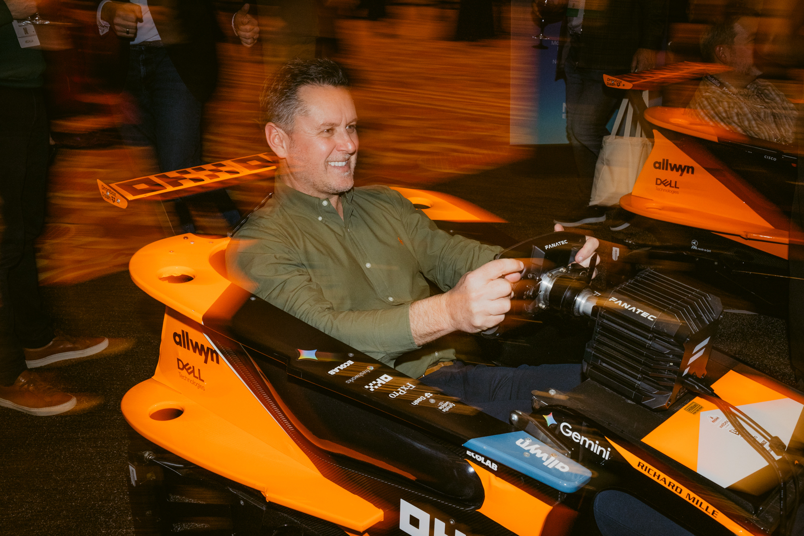 A man smiles while seated in an orange racing simulator car, holding the steering wheel. The background is slightly blurred, suggesting motion and a lively atmosphere, with other people and similar simulators visible.