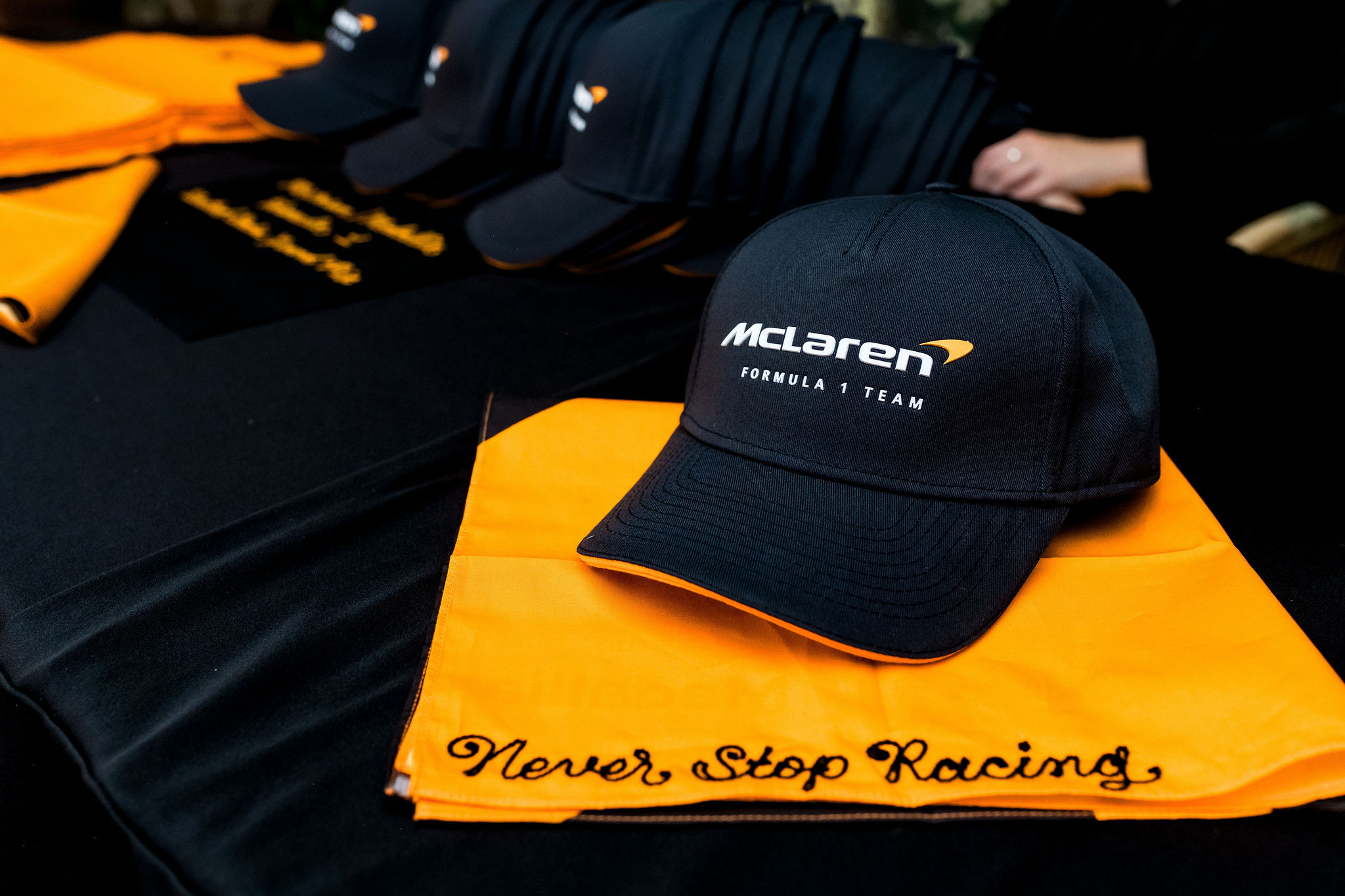 A black McLaren Formula 1 Team cap sits on an orange cloth with Never Stop Racing embroidered in black, with more caps and cloths arranged in the background on a black table.