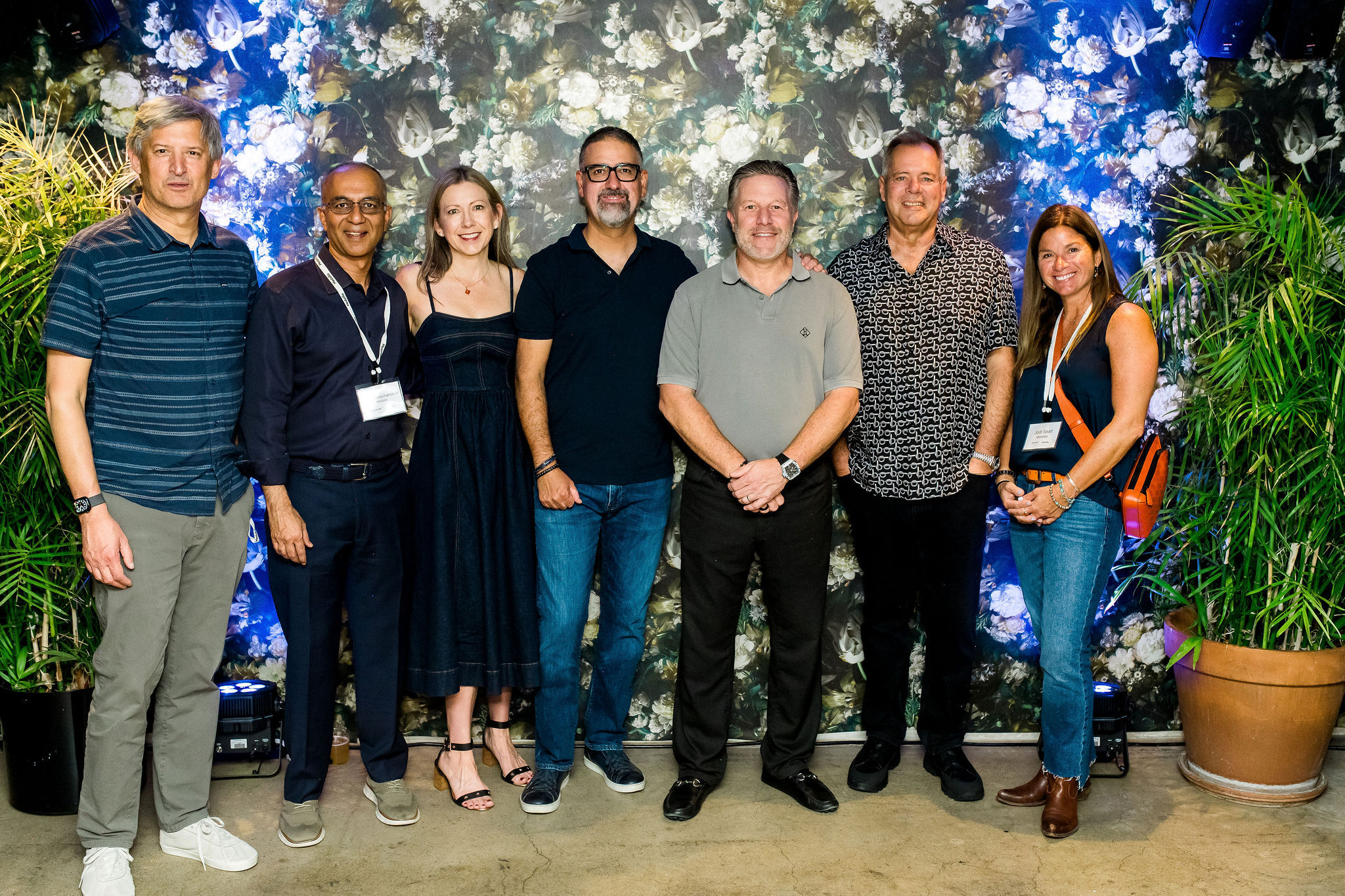 Seven people stand together smiling in front of a floral-patterned backdrop with green plants on each side. They are dressed in semi-casual attire and wearing conference name badges. The atmosphere appears friendly and informal.
