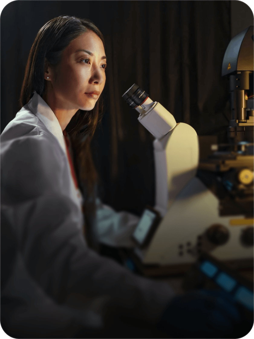 Una mujer con una bata blanca observa un microscopio en un laboratorio con poca luz, concentrada en su trabajo científico.