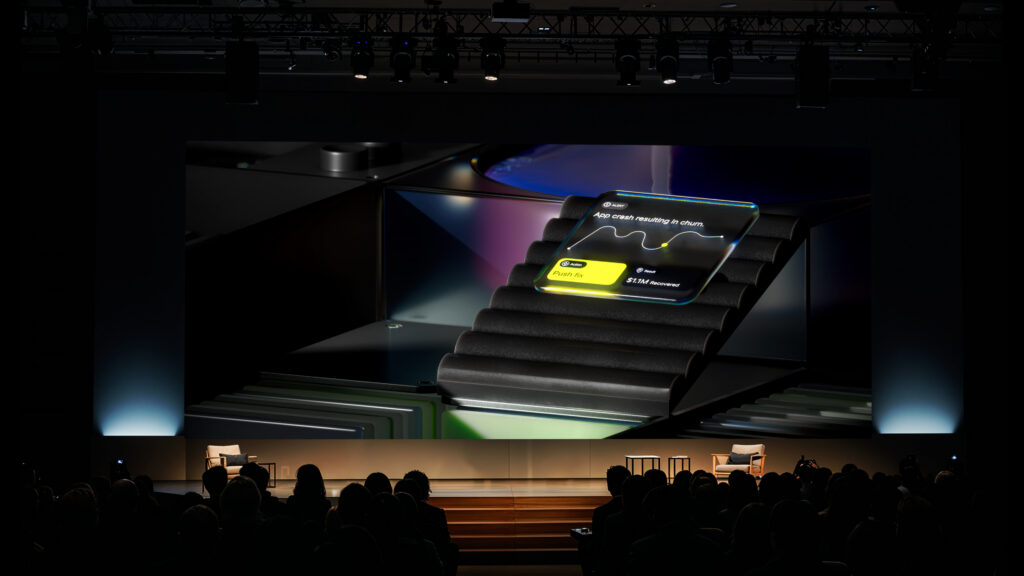 A large audience watches a stage presentation showing an enlarged smartwatch screen with a wellness app displaying heart rate data and a black band, projected onto a wide screen above the stage.