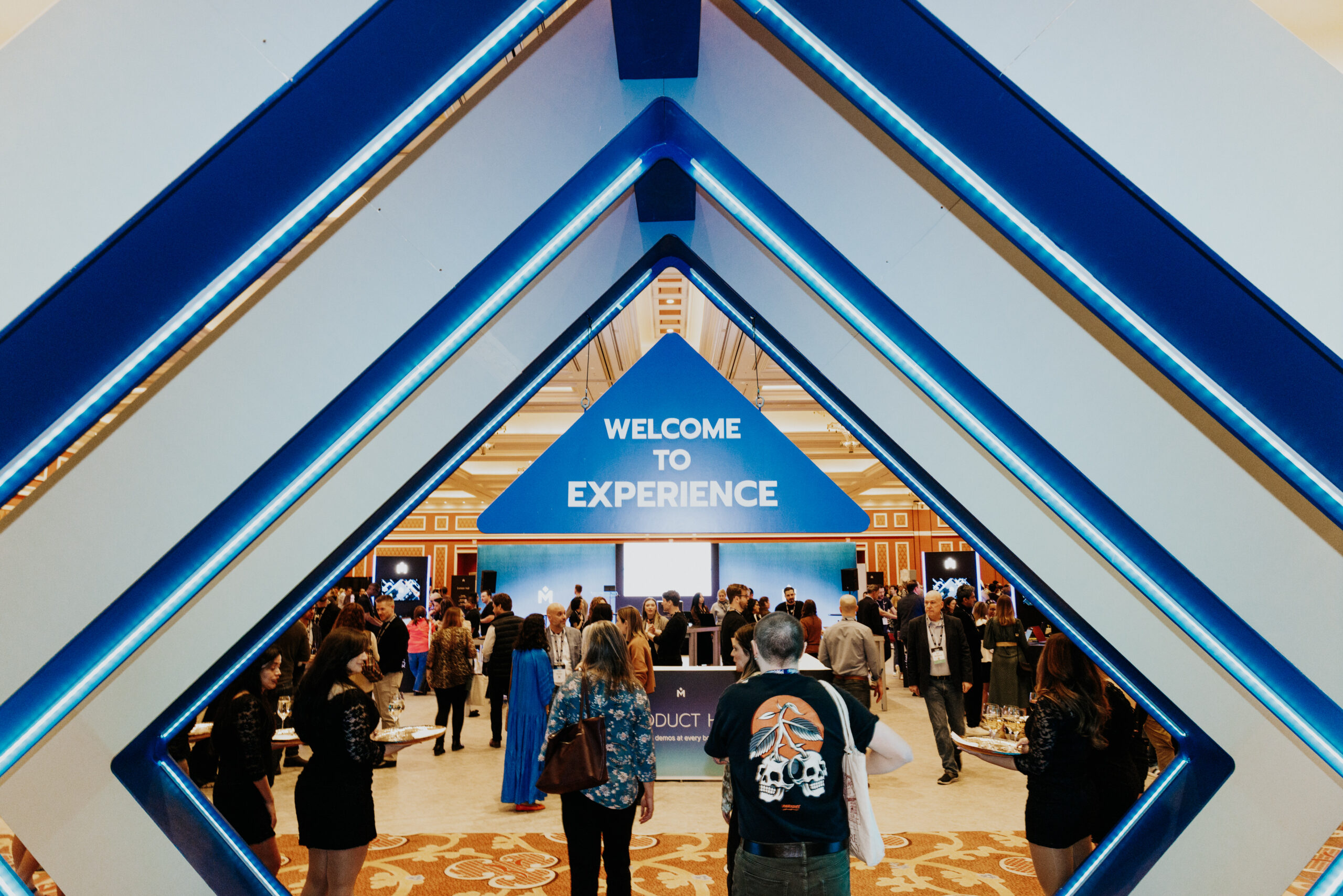 People enter a large event through a blue geometric archway. A sign inside reads WELCOME TO EXPERIENCE. Attendees gather and interact in a spacious, well-lit venue.