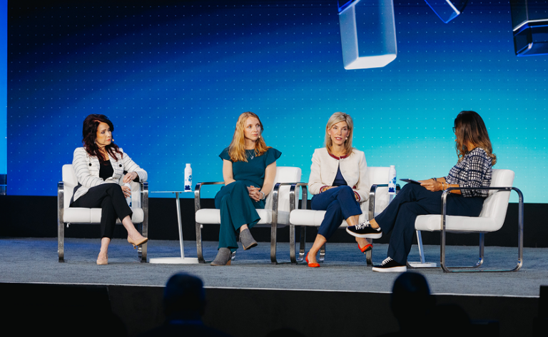 Four women sit on a stage in white chairs, engaged in a panel discussion. Blue background with geometric shapes and water bottles on tables are visible.