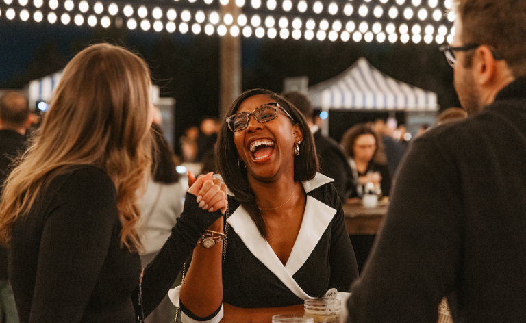 A woman wearing glasses laughs and holds hands with a friend at an outdoor event, surrounded by people and string lights, with striped tents in the background.