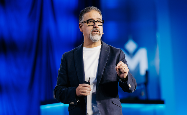 A man with glasses and a gray beard speaks onstage, wearing a dark blazer and white shirt, holding a small device, with a blue background and logo behind him.