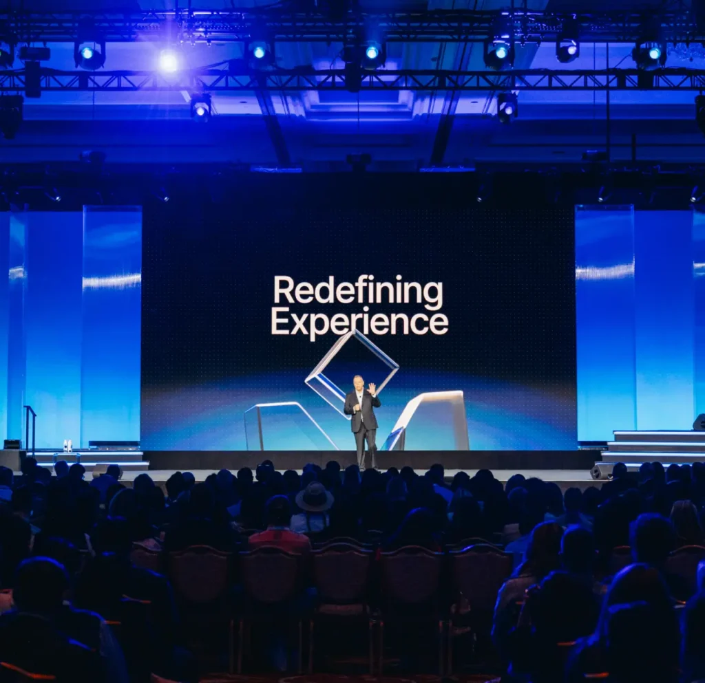 A speaker stands on stage in front of a large screen displaying the words Redefining Experience as an audience watches in a dimly lit conference hall with blue lighting.