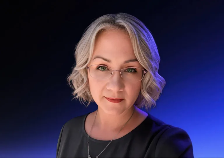 A woman with short, wavy blonde hair and glasses, wearing a black top and necklace, poses against a dark blue gradient background.