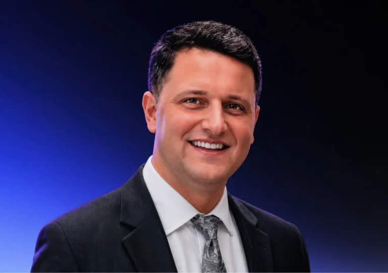 A man in a dark suit and patterned tie smiles at the camera, standing against a gradient blue and black background.