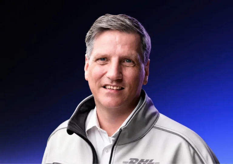 A man with short gray hair wearing a gray DHL jacket and white shirt smiles in front of a dark blue gradient background.