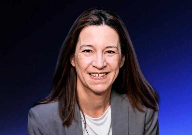 A woman with straight brown hair, wearing a gray blazer and white top, smiles at the camera with a blue gradient background behind her.
