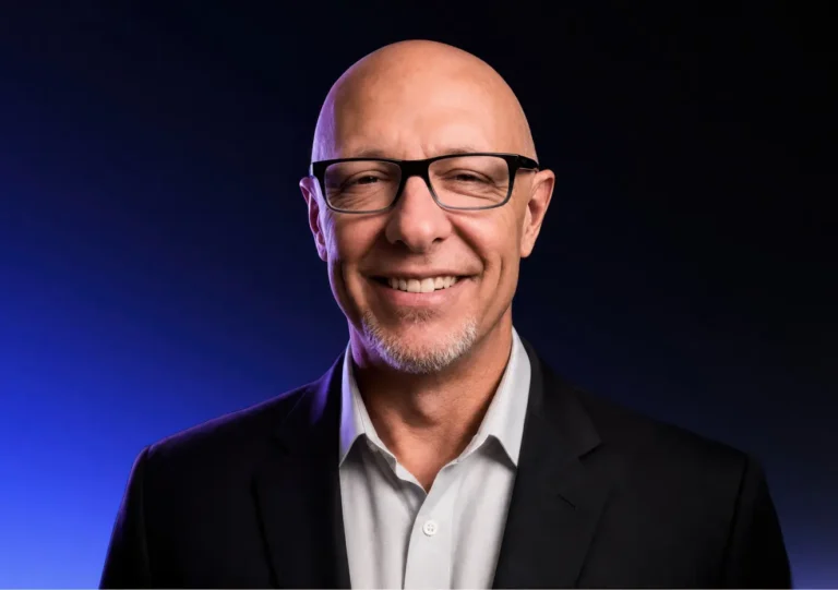 Bald man with glasses and a short beard, smiling and wearing a dark suit jacket over a light shirt, standing against a dark gradient background with blue and black tones.