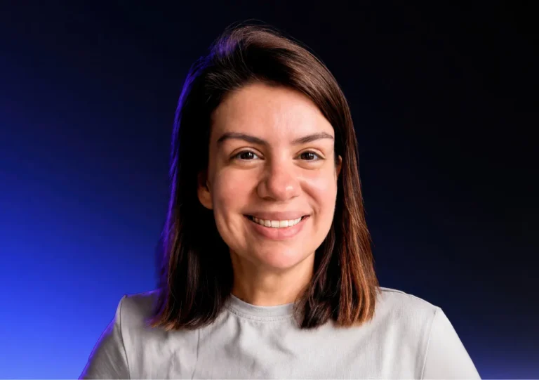 A woman with shoulder-length brown hair smiles at the camera. She is wearing a light gray top, and the background features a gradient from dark blue to black.