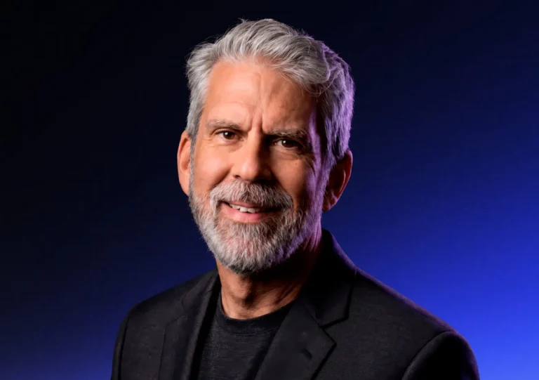 Smiling older man with gray hair and a beard wearing a black blazer and dark shirt, posed against a dark, blue-gradient background.