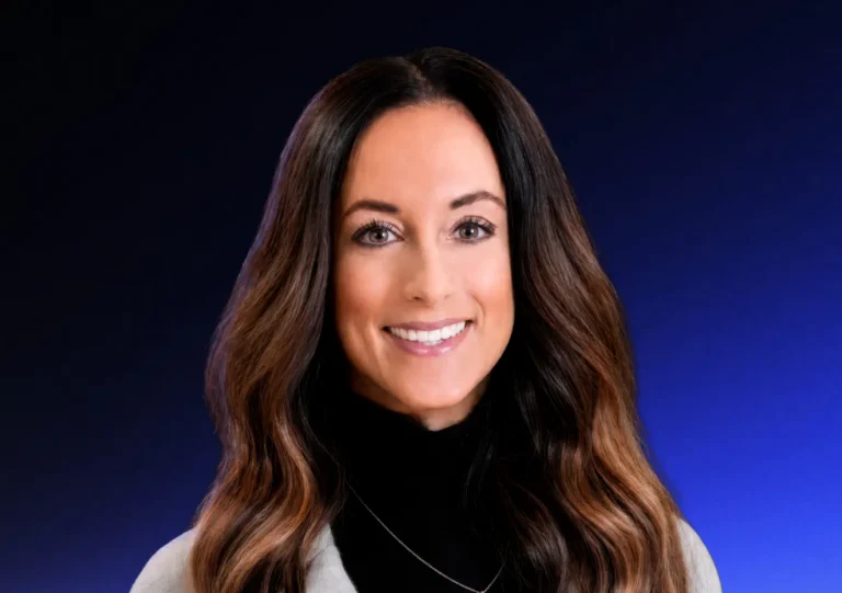 Woman with long wavy brown hair smiles at the camera, wearing a black turtleneck and light-colored blazer, against a blue gradient background.