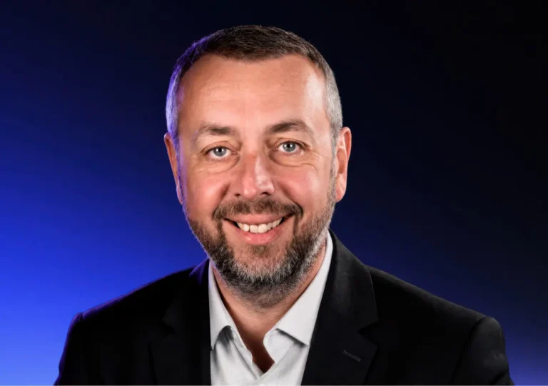 A smiling man with short hair and a beard wears a black blazer and light shirt, posing against a dark blue gradient background.