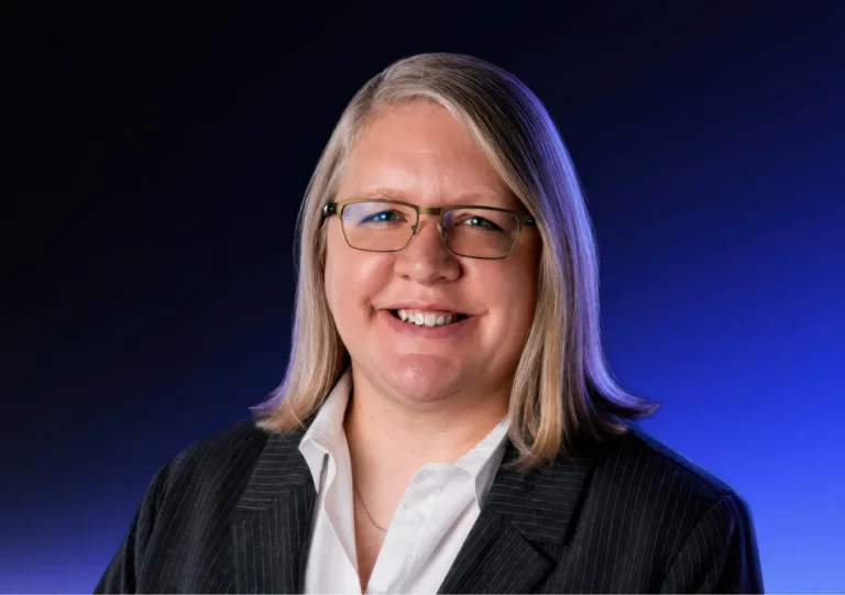 A person with shoulder-length blonde hair, glasses, and a pinstripe suit jacket over a white shirt smiles at the camera against a blue gradient background.