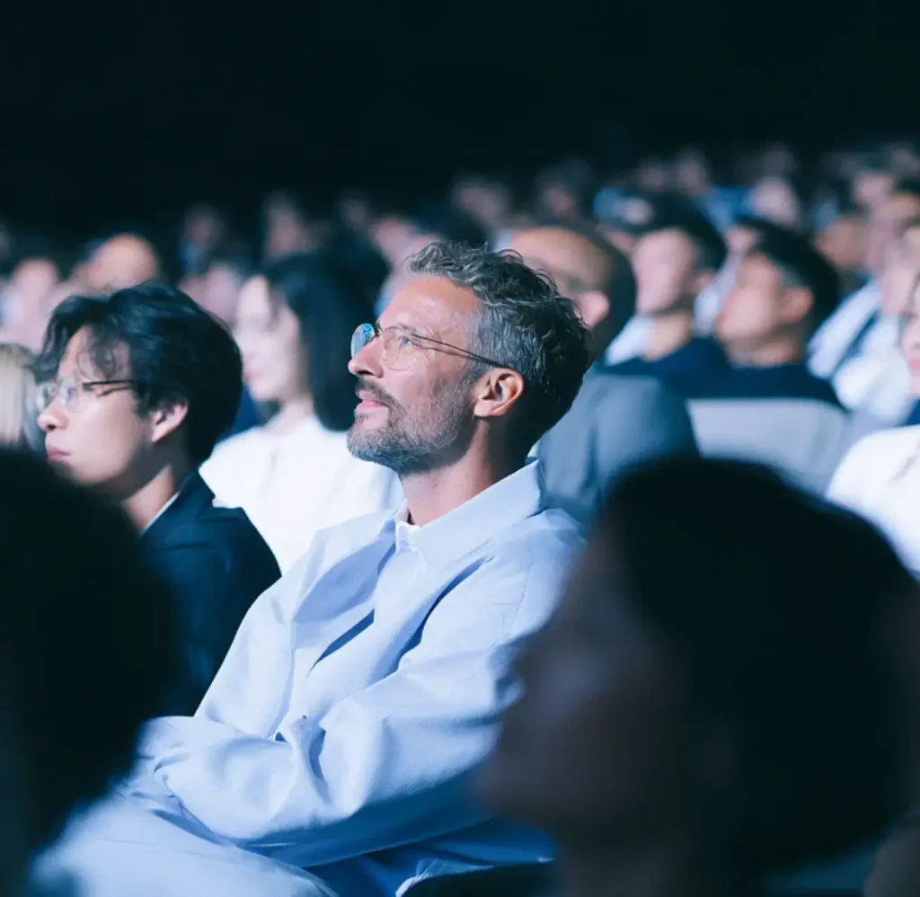 Un pubblico che assiste a una presentazione in una sala buia, con in primo piano un uomo con gli occhiali che guarda verso l’alto.
